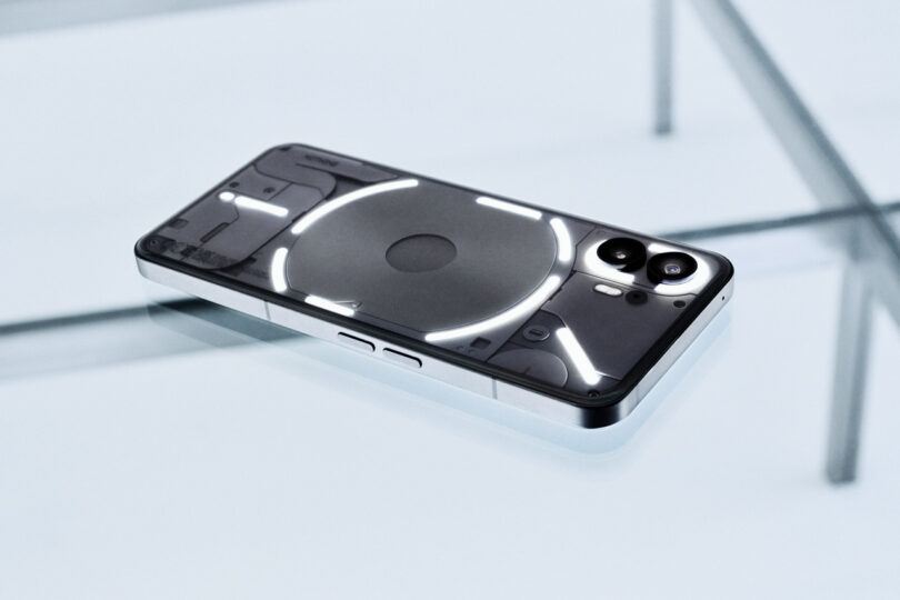 A smartphone with a transparent back, showing internal components and illuminated LED strips, rests on a glass table.