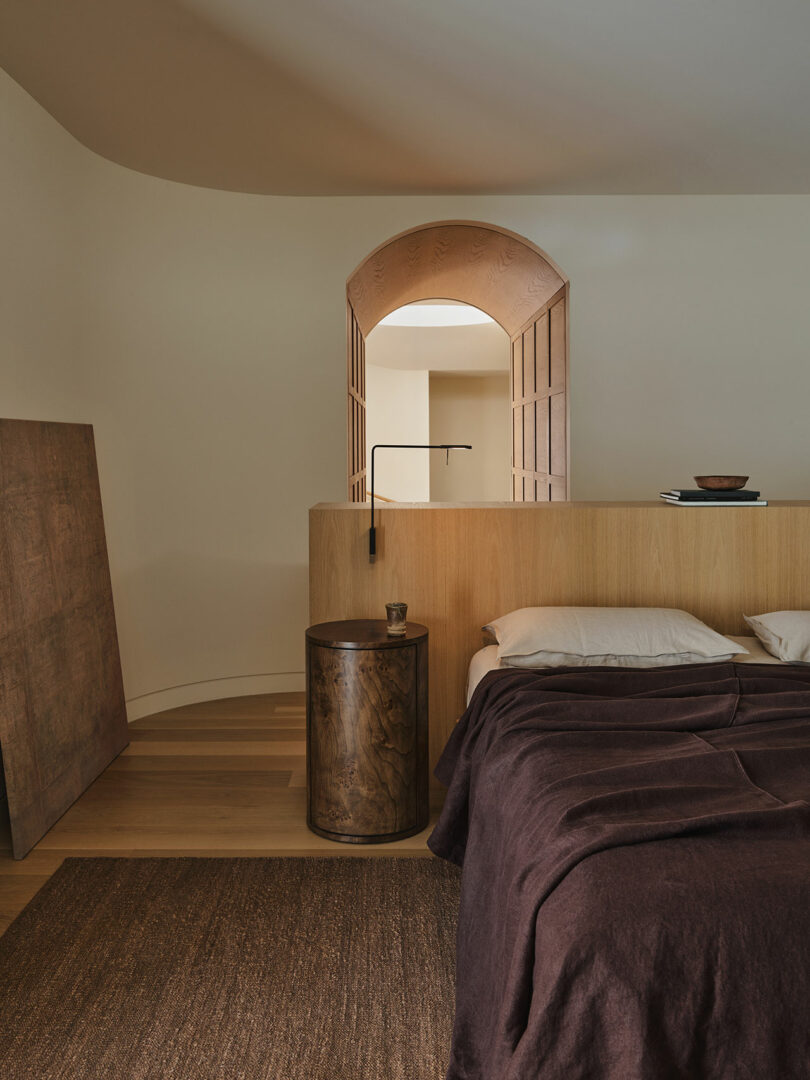 Minimalist bedroom with wooden accents, a brown blanket on the bed, a round wooden side table, and an arched doorway with folding doors in the background.