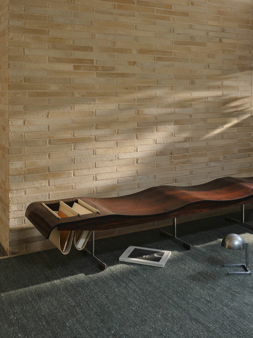 A modern wooden bench with built-in magazine storage sits against a beige brick wall; an open book and a metallic sphere are on the dark carpeted floor nearby.