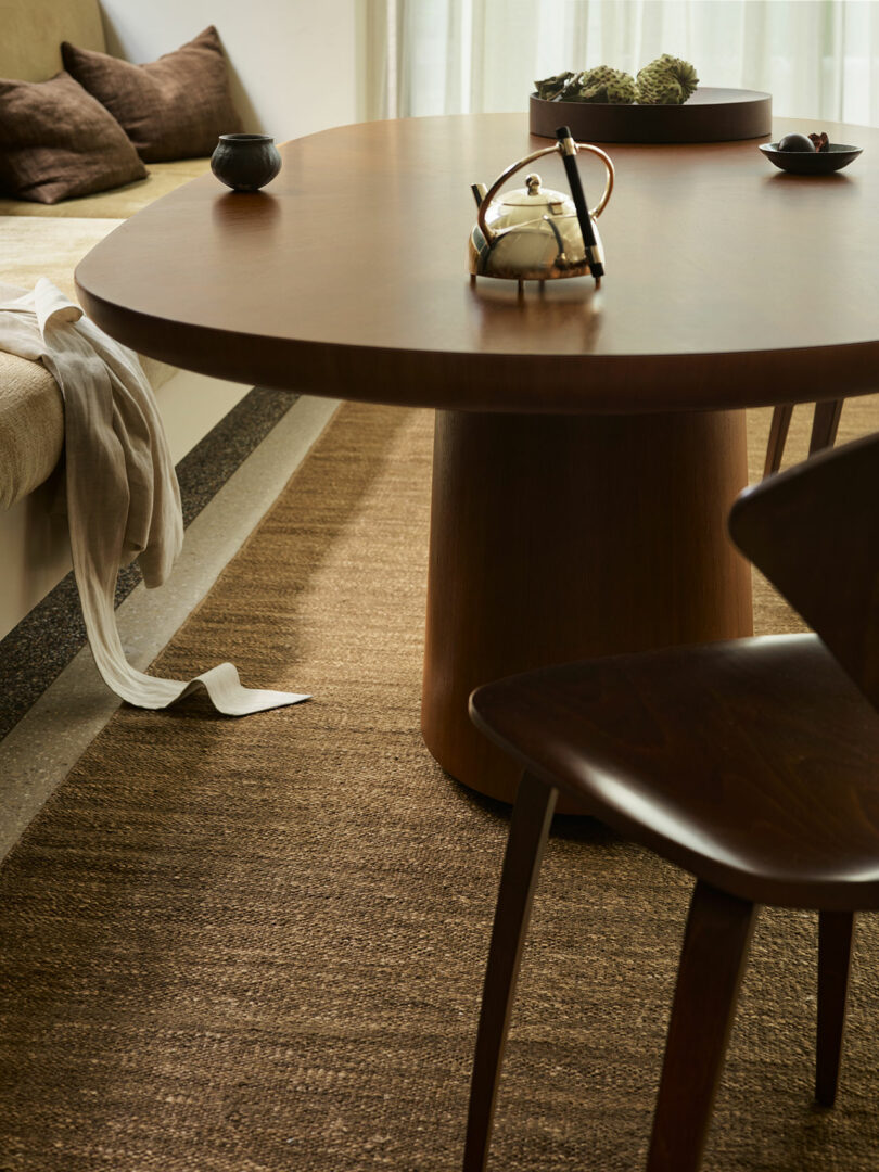 Round wooden table with a teapot and cup, beside a brown chair and bench with cushions, on a woven rug in a sunlit room.