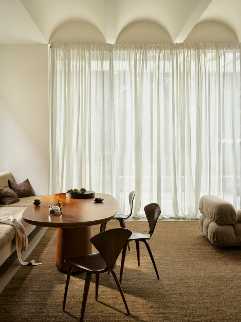 A modern dining area with a wooden table, three chairs, a beige cushioned bench, sheer floor-to-ceiling curtains, and a neutral-toned carpet.