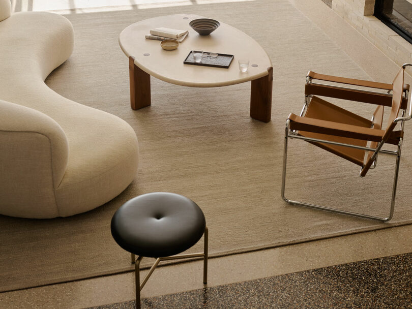 A modern living room with a curved beige sofa, a light wood coffee table with books and bowls, a leather and chrome chair, and a round black stool on a beige rug.