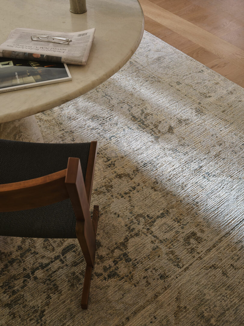 A round marble table with a newspaper and magazine on top, a wooden chair with a dark seat, and a patterned rug on a wooden floor.