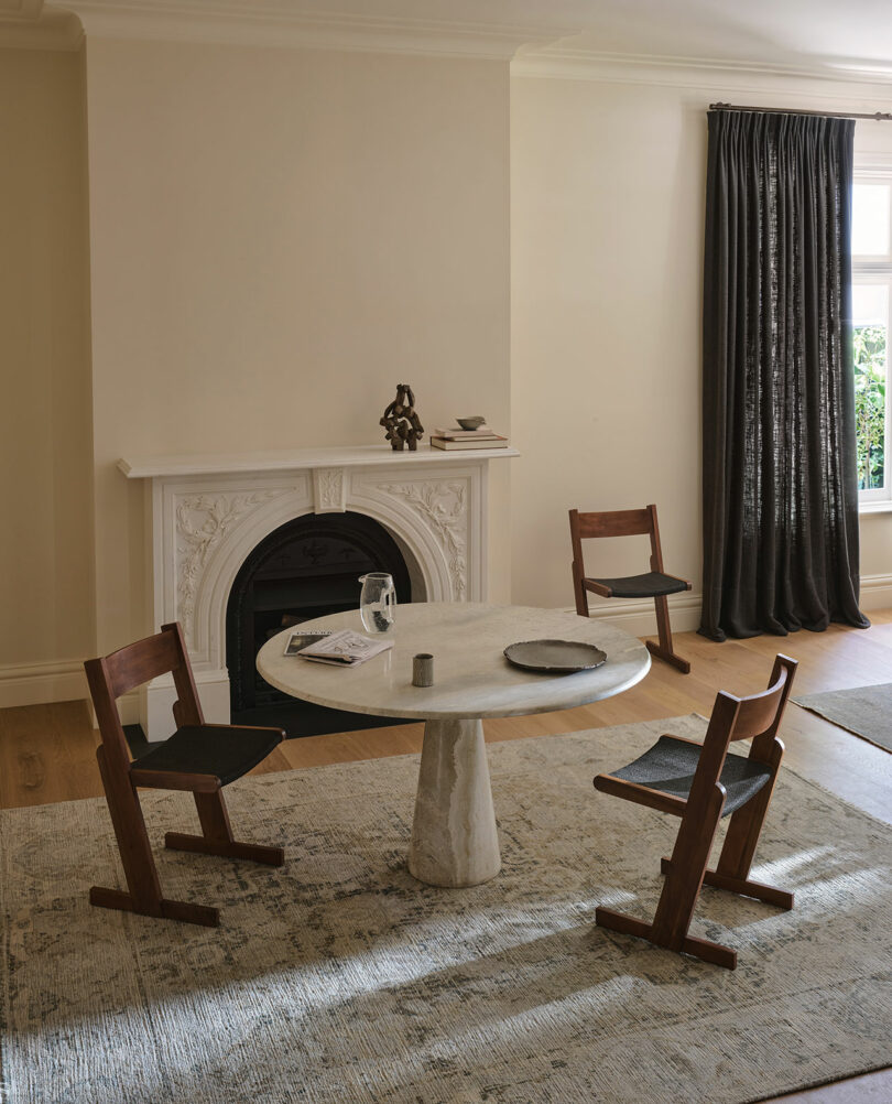 A round marble table with three wooden chairs on a patterned rug in front of a white decorative fireplace in a sunlit room with dark curtains.