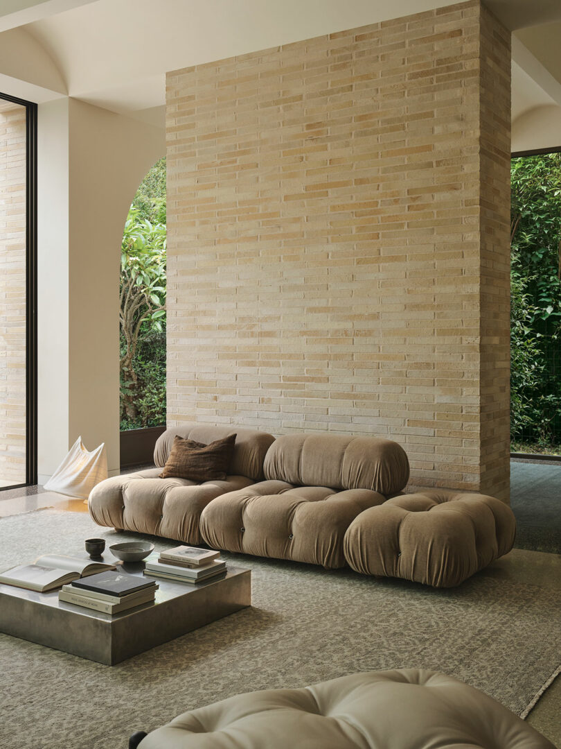 A modern living room with a tufted beige sofa, a low coffee table with books and cups, a textured rug, and floor-to-ceiling windows showing greenery outside.