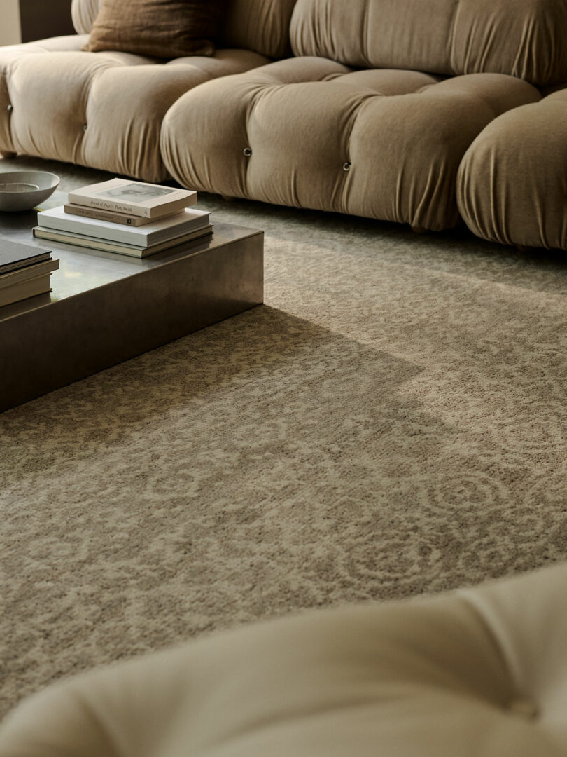 A beige tufted sofa sits next to a low, rectangular coffee table with books on top, placed on a patterned neutral carpet in a softly lit living room.