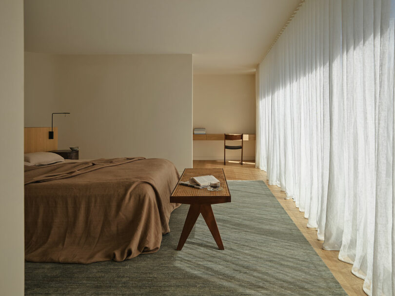 Minimalist bedroom with a brown bedspread, wooden furniture, a small desk and chair, light wood floors, and large sheer curtains letting in natural light.
