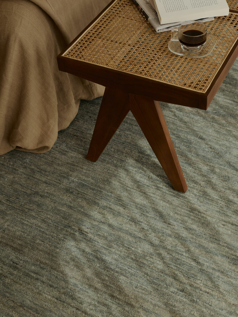 A wooden side table with a woven top holds an open book and a glass cup of coffee, next to a bed with a beige blanket on a textured gray rug.