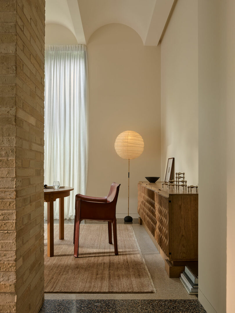 A minimalist room with a wooden table, a brown chair, a floor lamp with a round paper shade, a wooden sideboard, stacked books, and sheer curtains letting in natural light.