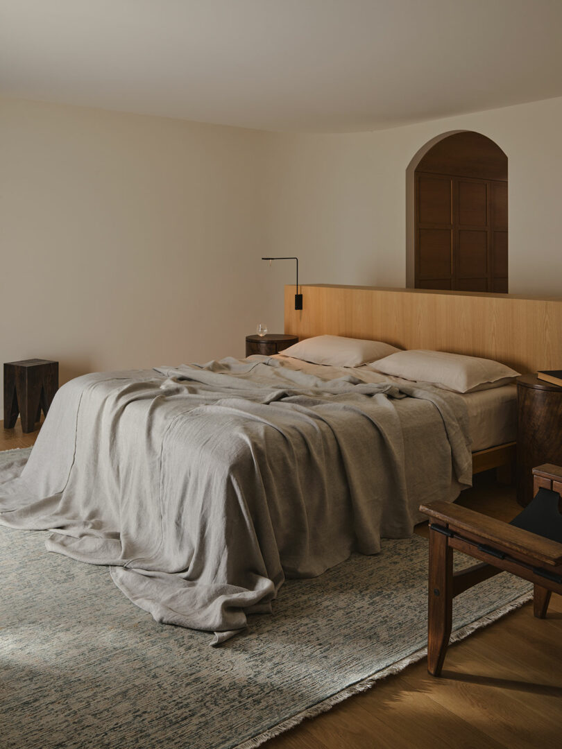 A modern bedroom with a wooden headboard, unmade bed with beige linens, two nightstands, a rug, and minimal decor.