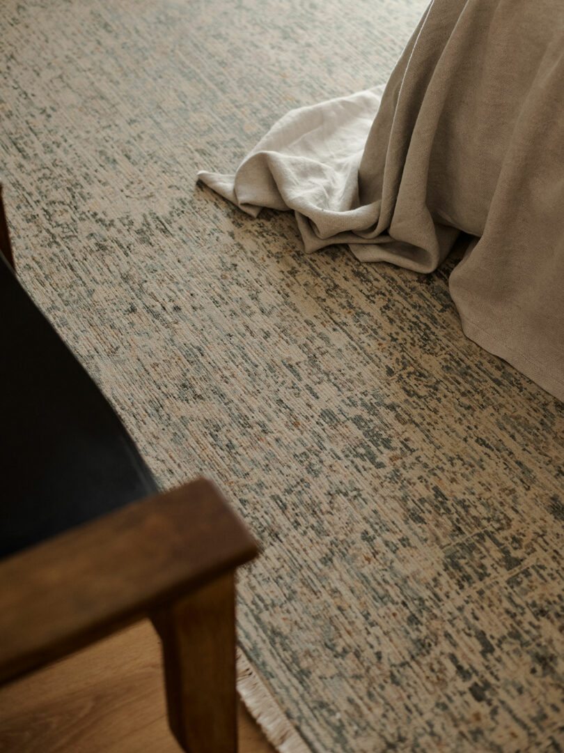A close-up of a patterned rug with a wooden chair in the corner and a light-colored fabric draped over the edge of a surface.