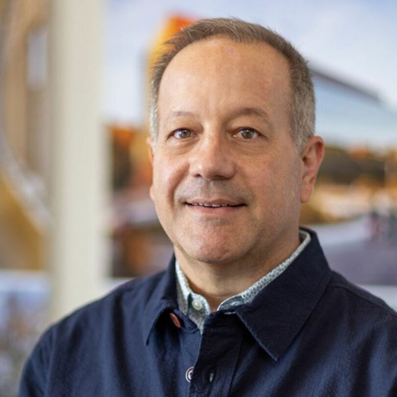 Barry Ginder, a middle-aged man in a navy blue jacket and light patterned shirt, stands indoors with blurred artwork or photographs visible in the background.