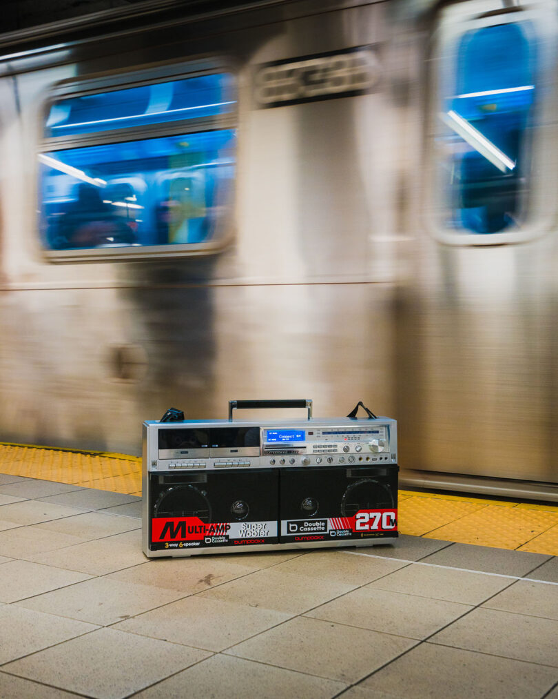 A portable boombox sits on a subway platform with a blurred train passing by in the background.