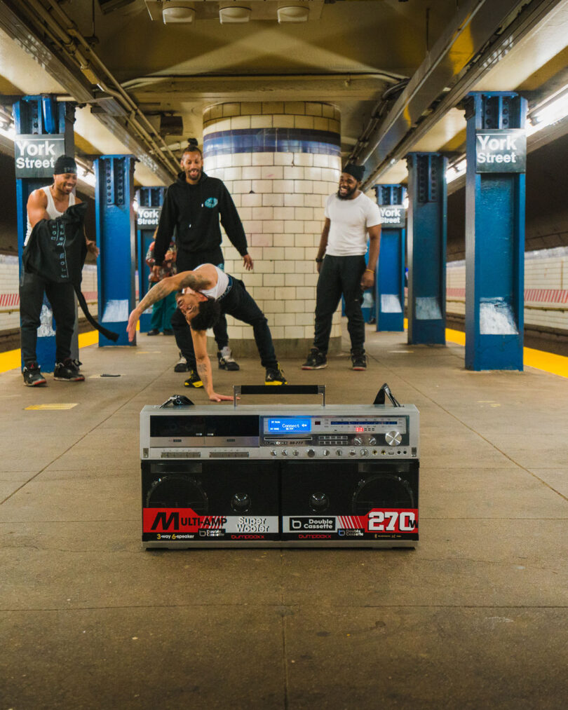 Four men perform and watch a breakdancer in a subway station with a large boombox placed in the foreground.
