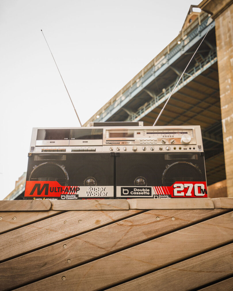 A vintage portable boombox with antennas and dual cassette decks sits on a wooden surface outdoors, with part of a bridge visible in the background.
