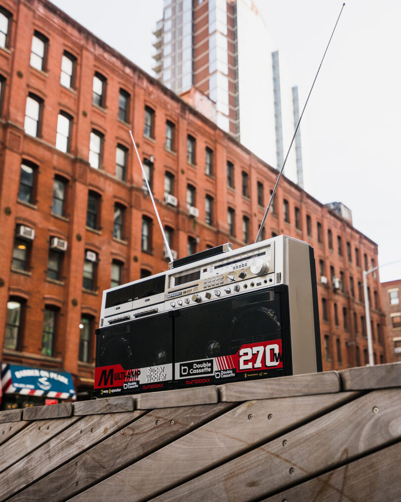 A vintage boombox with extended antennas sits on a wooden surface outdoors, with brick apartment buildings in the background.
