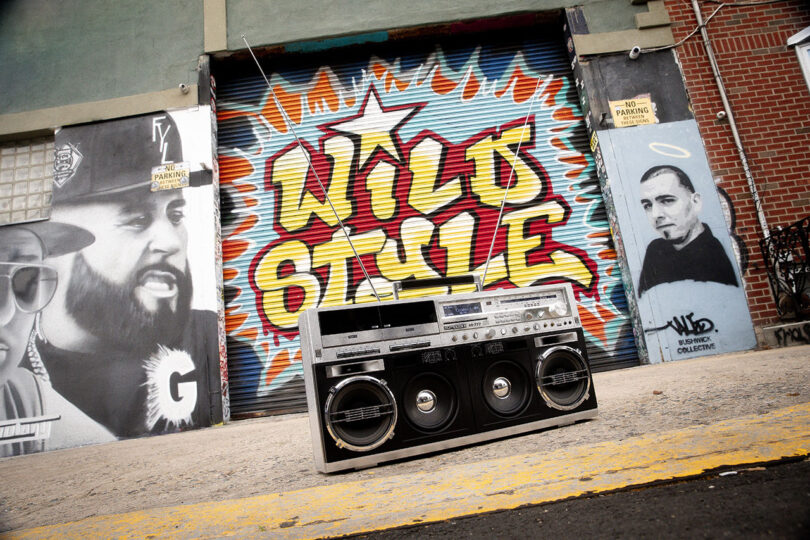 A large boombox sits on the ground in front of a mural reading "WILD STYLE," surrounded by black-and-white portraits and graffiti art on a city wall.