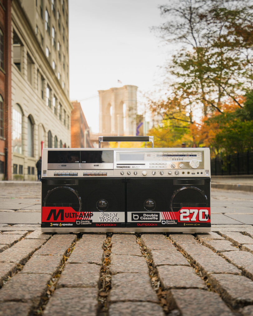 A large vintage boombox sits on a cobblestone street with a bridge and buildings visible in the background.