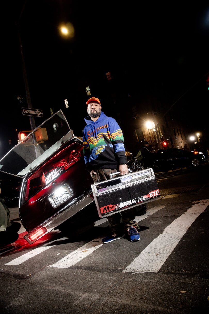 A man in a colorful jacket stands on a city street at night holding a metal case, with an open car door and streetlights in the background.