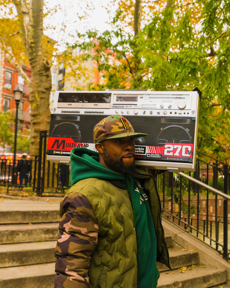 A man wearing a camouflage jacket and hat carries a large vintage boombox on his shoulder in an outdoor park setting with autumn trees.
