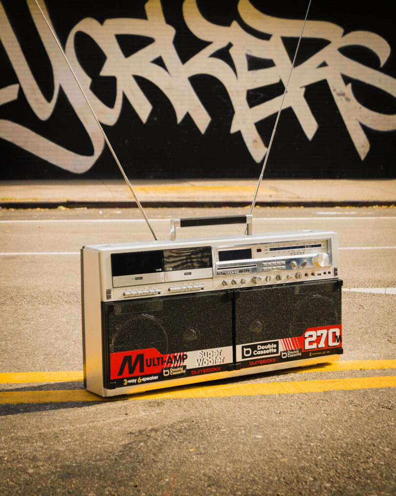 A vintage boombox with extended antennas sits on a street’s yellow line, with a large graffiti-covered wall in the background.