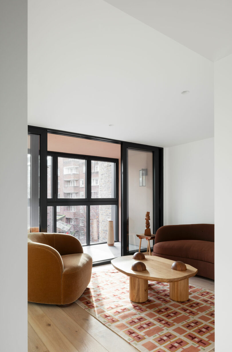 Modern living room with a brown sofa, tan armchair, geometric rug, wooden coffee table, and large floor-to-ceiling windows overlooking an urban view.