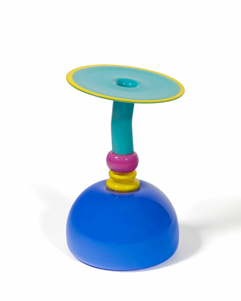 Colorful ceramic object with a wide blue base, pink and yellow rings, teal stem, and a flat turquoise top, resembling an abstract or modern candlestick holder.