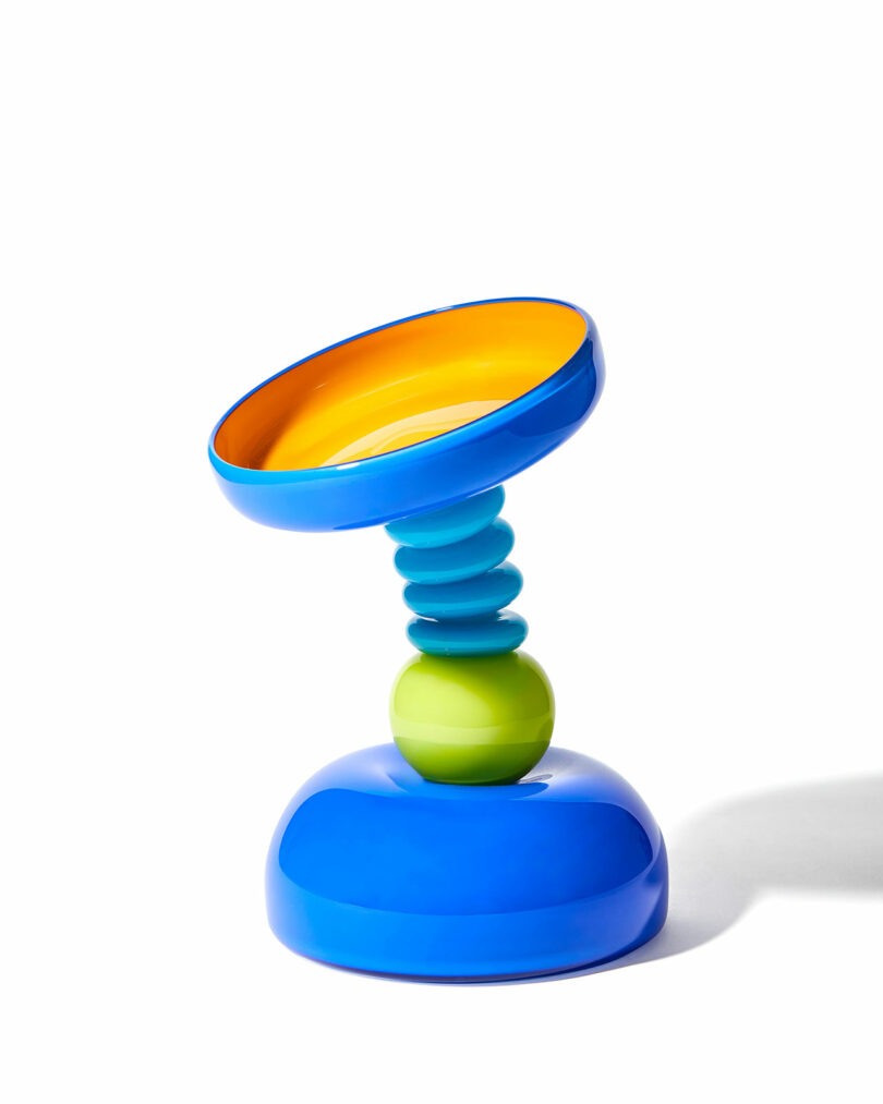 A colorful, modern abstract sculpture with a blue base, a green orb, stacked blue discs, and a tilted, circular yellow and blue top, set against a white background.