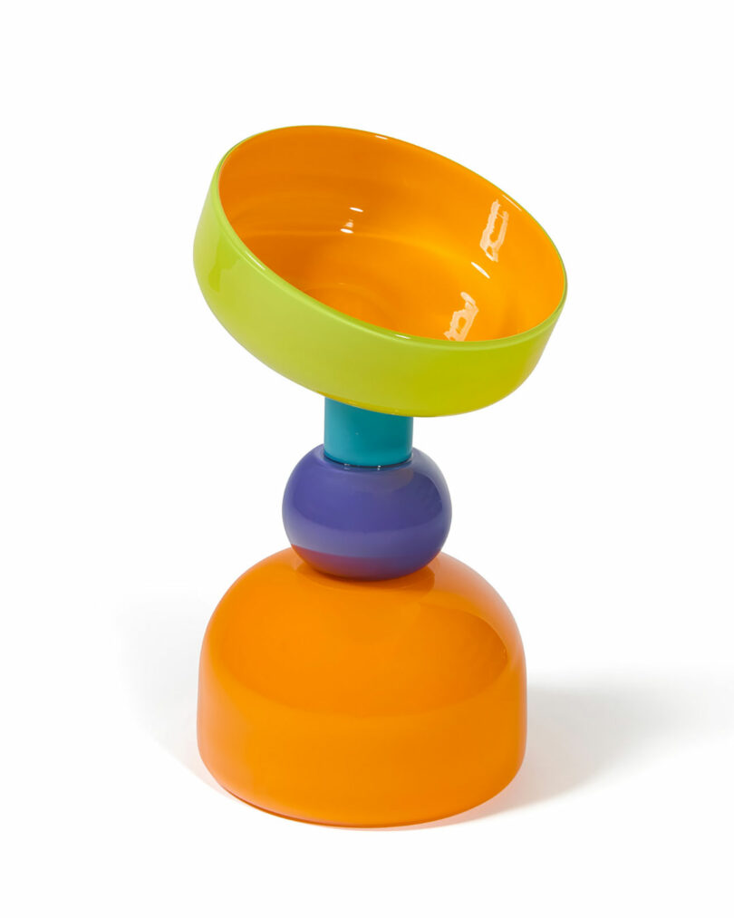 A colorful, abstract object featuring a tilted green and orange bowl on top of stacked purple, blue, and orange rounded shapes.