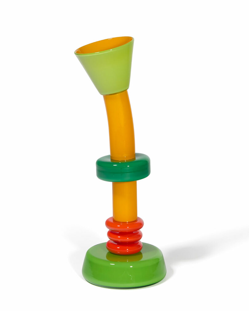 A colorful glass object with a green base, red and green rings, an orange vertical section, and a green flared top, set against a white background.