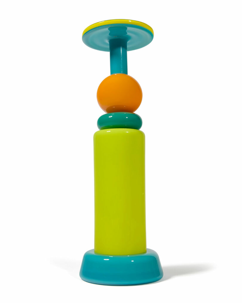 A colorful Cedric Mitchell JOOPITER pedestal with a bright yellow-green base, turquoise accents, and an orange sphere in the center, photographed against a white background.