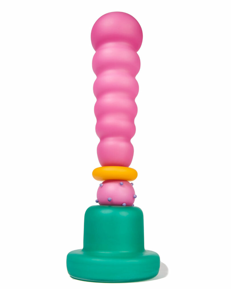 A colorful abstract sculpture inspired by Cedric Mitchell JOOPITER, featuring a tall, rounded pink column with bulbous segments, perched on a green base with yellow and purple accents.