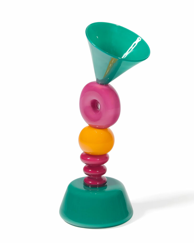 A colorful abstract glass sculpture with stacked geometric shapes in teal, pink, yellow, and orange, featuring a cone, disks, and a rounded base.