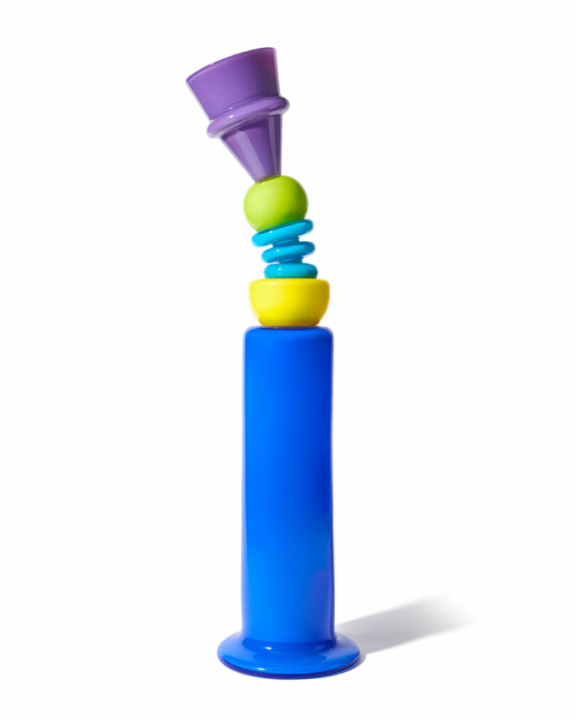 A colorful abstract sculpture by Cedric Mitchell JOOPITER features stacked geometric shapes in purple, green, turquoise, yellow, and blue, standing boldly on a white background.