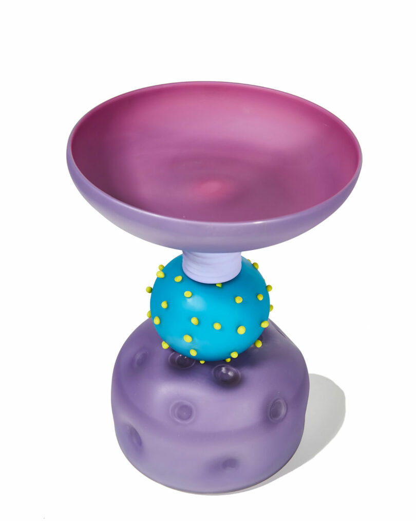 A colorful, sculptural glass vessel by Cedric Mitchell JOOPITER features a wide purple-pink bowl, a blue sphere with yellow dots, and a purple textured base, all set against a white background.