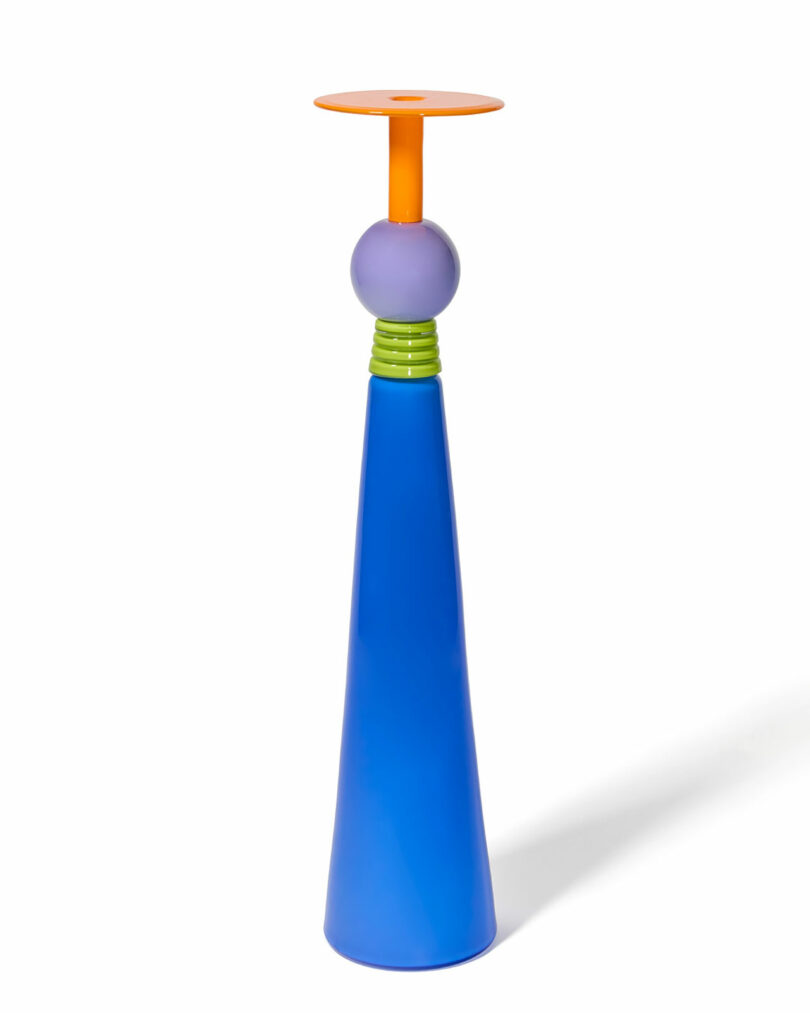 A tall, cone-shaped candle holder by Cedric Mitchell JOOPITER features a blue base, green rings, a purple sphere, and an orange circular top set against a white background.
