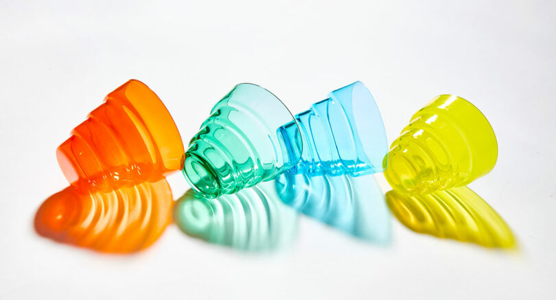 Four translucent, colored plastic cups—orange, green, blue, and yellow—are tipped on their sides in a row, casting matching shadows on a white surface.