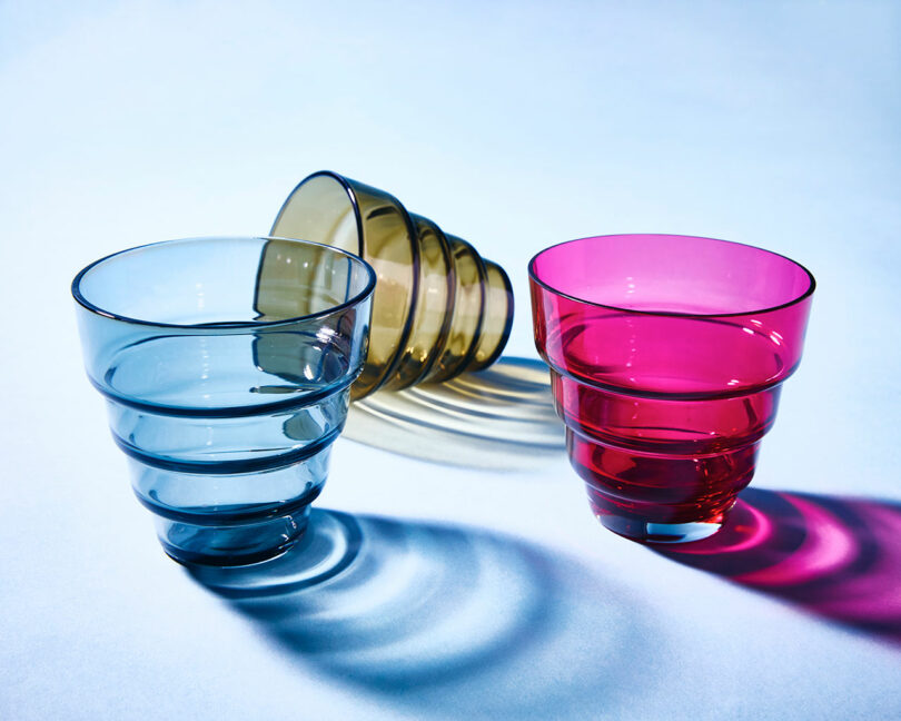 Three ribbed glass cups in blue, amber, and pink are arranged on a light surface, with the amber cup lying on its side and the others standing upright, casting colored shadows.