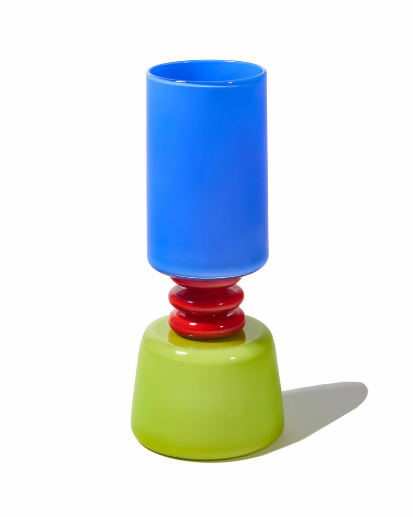 A colorful glass vase with a tall blue cylindrical top, red ringed middle, and a wide green base, set against a white background.
