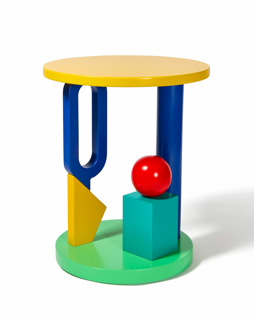 Colorful, geometric side table with a yellow top, blue and green cylindrical legs, a red sphere, and various angular shapes on a round green base.