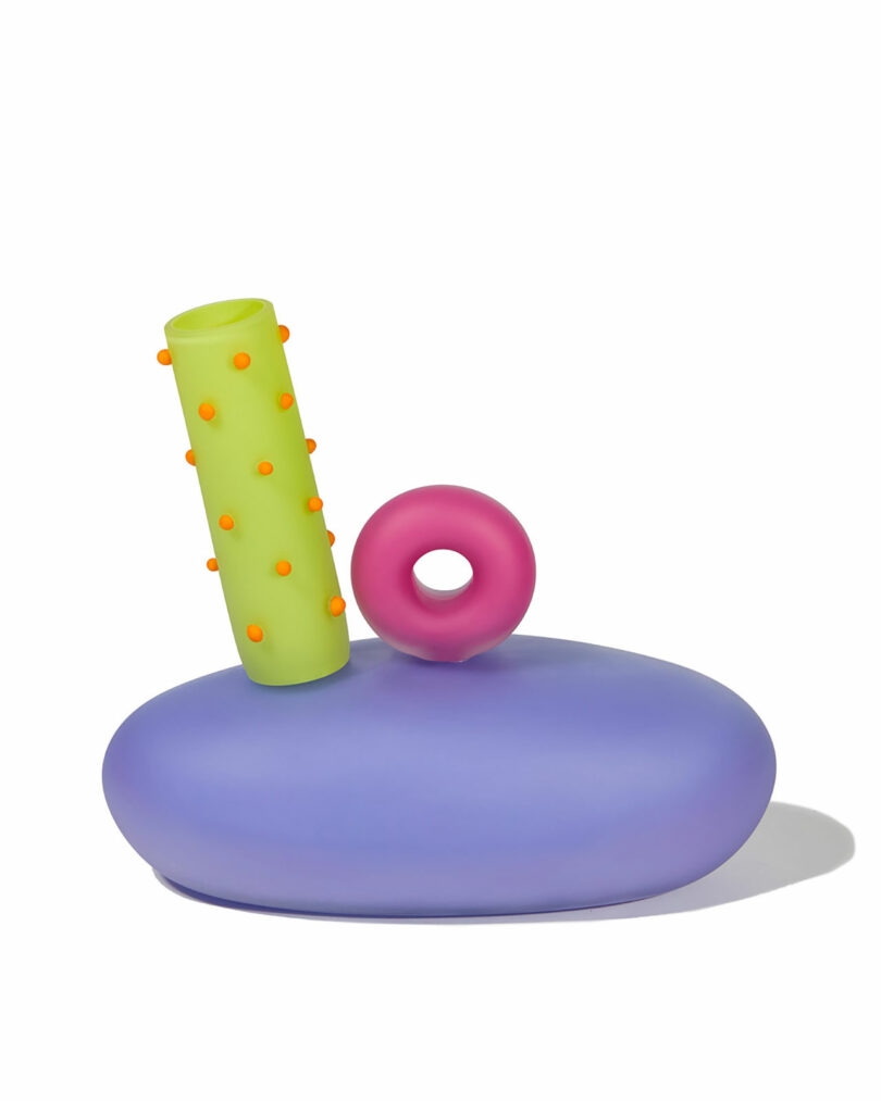 A sculptural object with a smooth purple oval base, a vertical green tube with orange dots, and a pink ring on top, all on a white background.