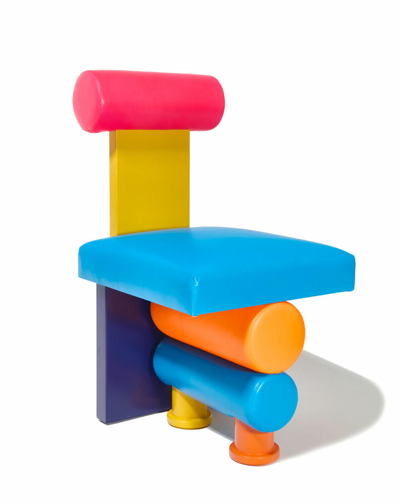 A chair with a geometric design, featuring a pink cylindrical backrest, yellow vertical support, blue seat, and orange and blue cylindrical legs, all in bright, solid colors.