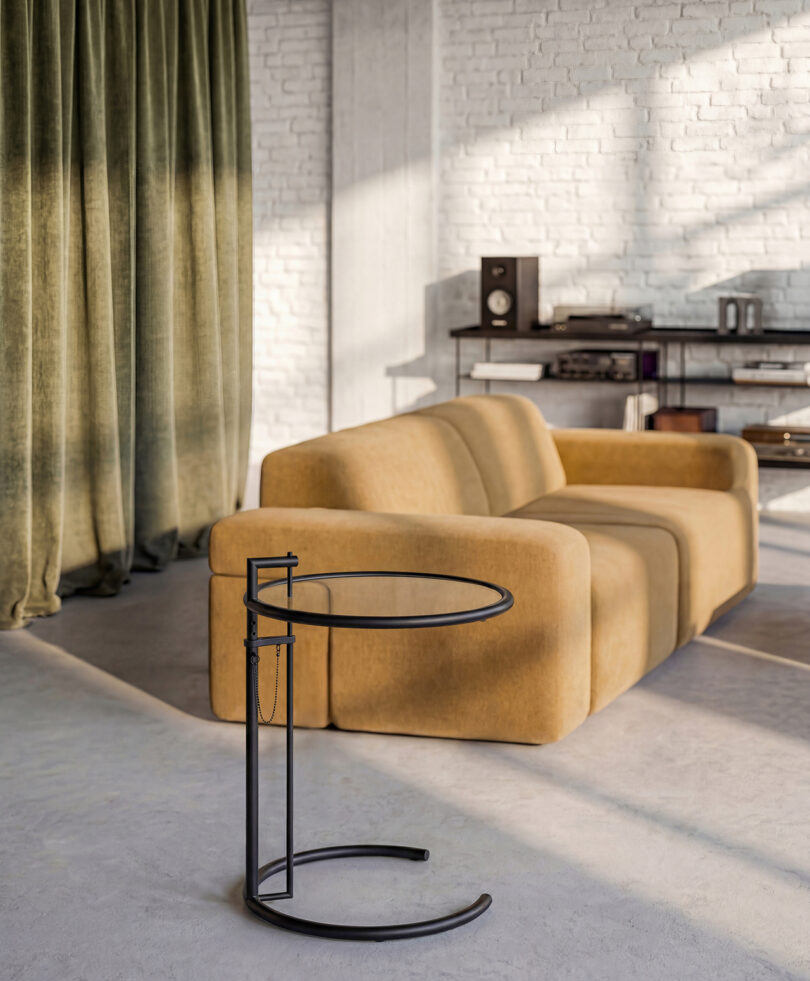 A modern living room designed by Gabriel Tan, featuring a mustard yellow sofa, a round glass side table, green curtains, and a white brick wall with shelves holding audio equipment.