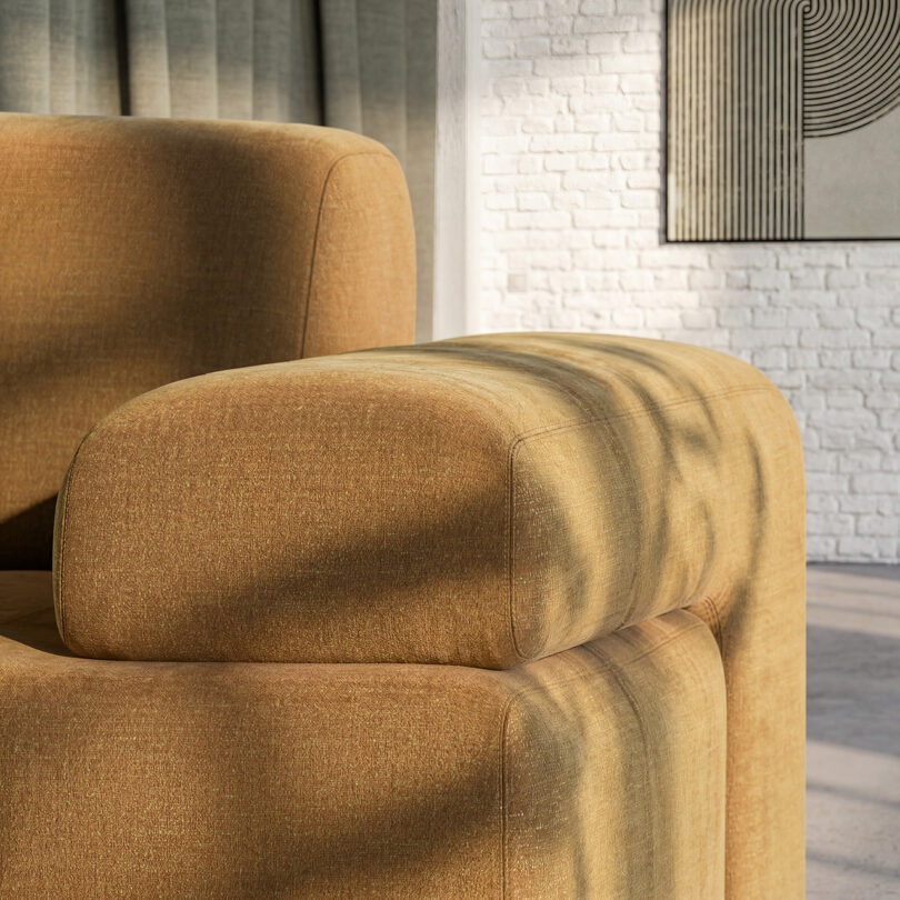 Close-up of a modern Gabriel Tan mustard-colored sofa with rounded edges, sunlight casting shadows, and part of a white brick wall and abstract artwork in the background.