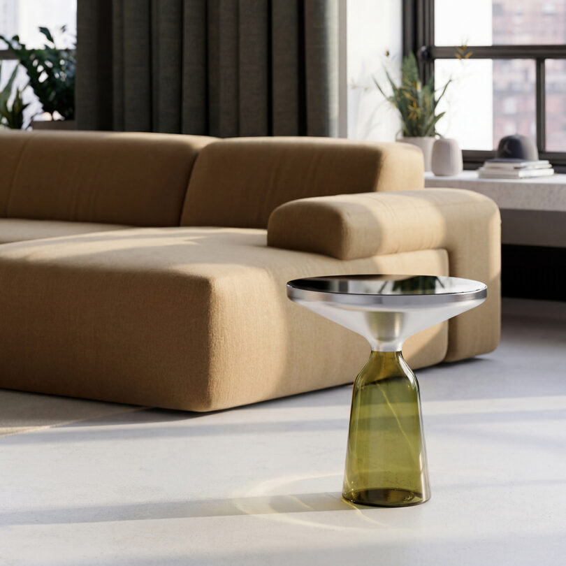 A modern living room with a beige sectional sofa, a Gabriel Tan green glass and metal side table, large windows, and neutral-colored curtains.