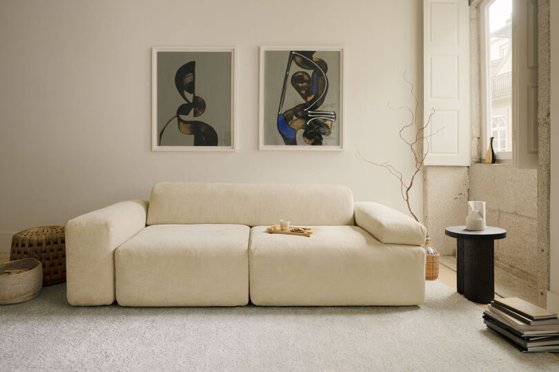A beige modern sofa by Gabriel Tan sits against a light wall with two abstract paintings above it; a side table holds a white pitcher and cups next to a window with natural light.