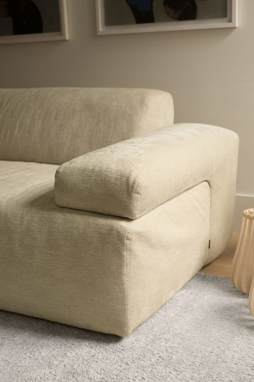 Close-up of the armrest and cushion of a light beige fabric sofa on a light gray rug, with part of a wooden table and framed artwork visible in the background, showcasing Gabriel Tan’s refined design aesthetic.