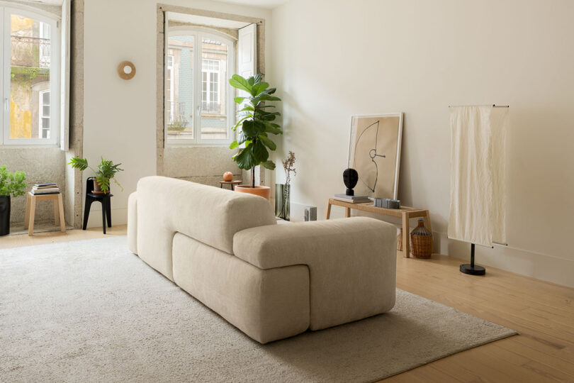 Minimalist living room with a Gabriel Tan beige sofa, potted plants, light wood floor, large windows, and modern decor accents.