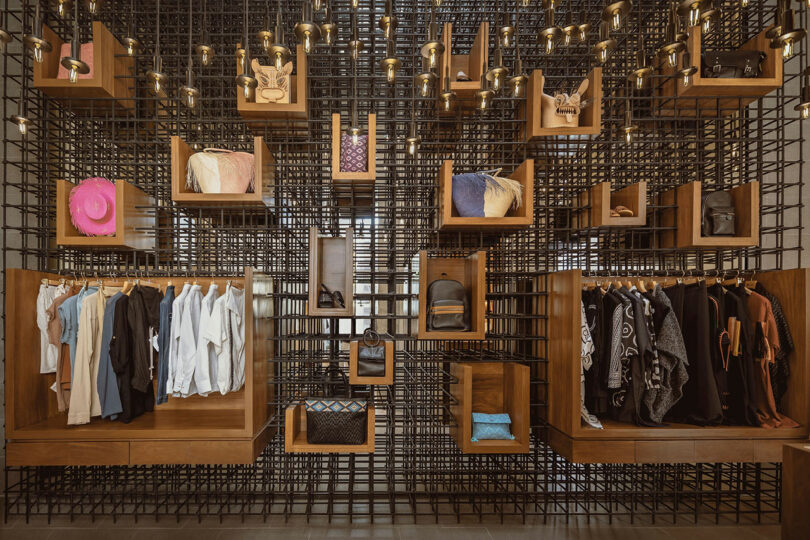 A retail display features clothing and accessories arranged in wooden boxes on a geometric black grid wall, with hanging lights above.