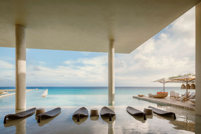Modern outdoor seating area with a submerged bar, infinity pool, lounge chairs, and umbrellas overlooking the ocean under a partly cloudy sky.